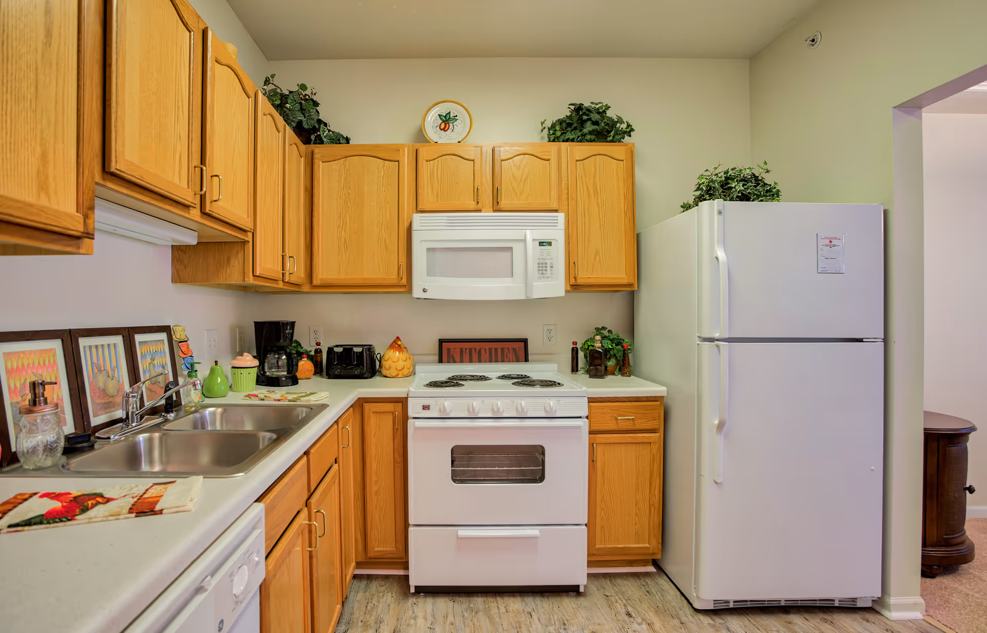 A kitchen with wooden cabinets, a white electric stove with oven, a white microwave above the stove, a white refrigerator, and a double sink. The countertop has various items including a coffee maker, toaster, soap dispenser, and decorative plants. The floor is wood-patterned and the walls are light-colored.