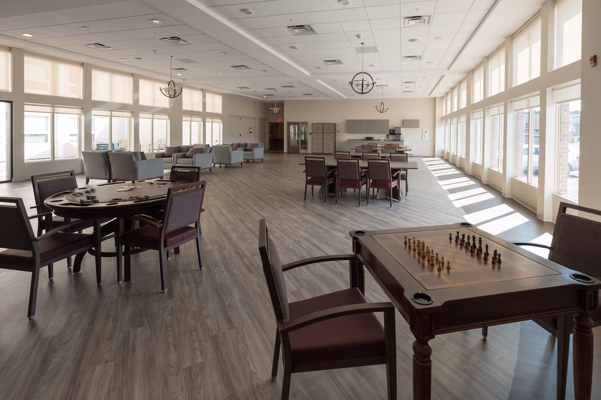 A spacious, sunlit senior living common room with game tables, chairs, and seating areas next to large windows.