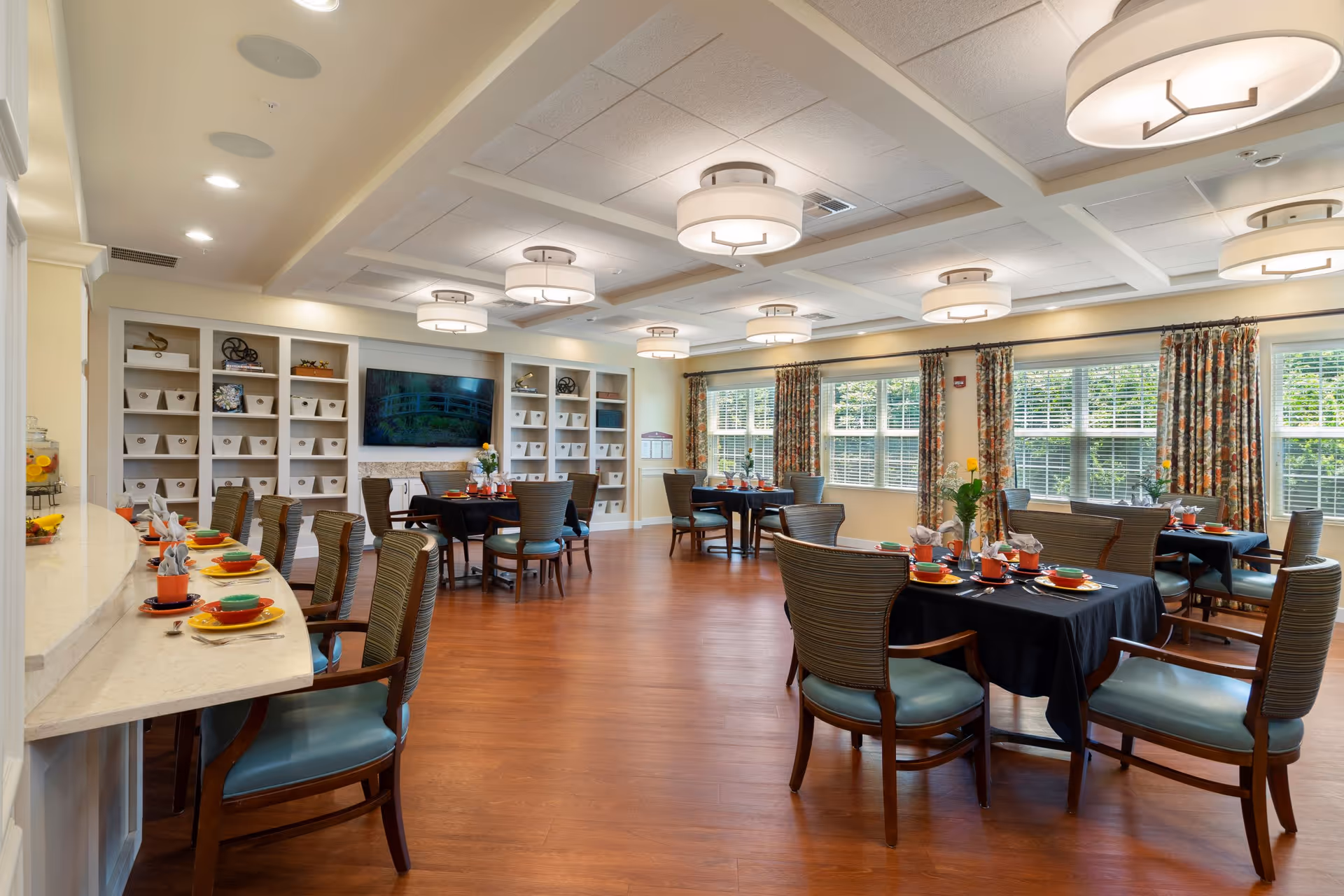 A bright and spacious dining room with multiple tables set for meals. The room features wooden floors, large windows with floral curtains allowing natural light, and modern ceiling lights. There is a long counter with chairs on the left side and built-in shelves with decorative items and a large flat-screen TV on the far wall.