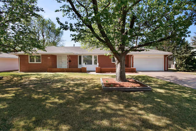 Single-story brick house with a large front yard featuring a mature tree in a mulched bed. The house has a white front door, a large window, and an attached two-car garage. The lawn is well-maintained and there are other trees and houses visible in the background under a clear blue sky.