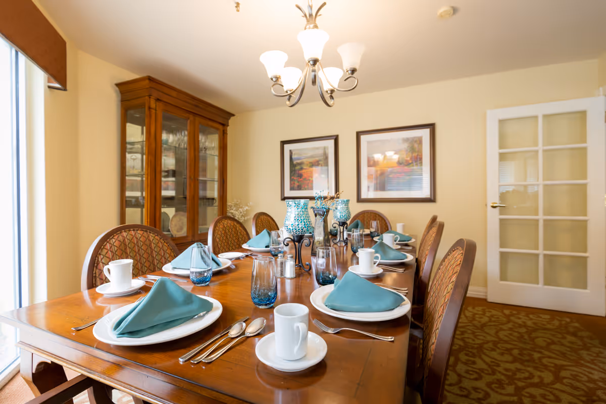 A dining room with a wooden table set for eight people. Each place setting includes a white plate, teal folded napkin, silverware, a blue-tinted glass, and a white coffee cup with a saucer. The room features a wooden china cabinet with glass doors, two framed landscape paintings on the wall, a chandelier with five lights, and a glass-paneled door. The carpet has a patterned design, and there is a window with a brown valance on the left side.