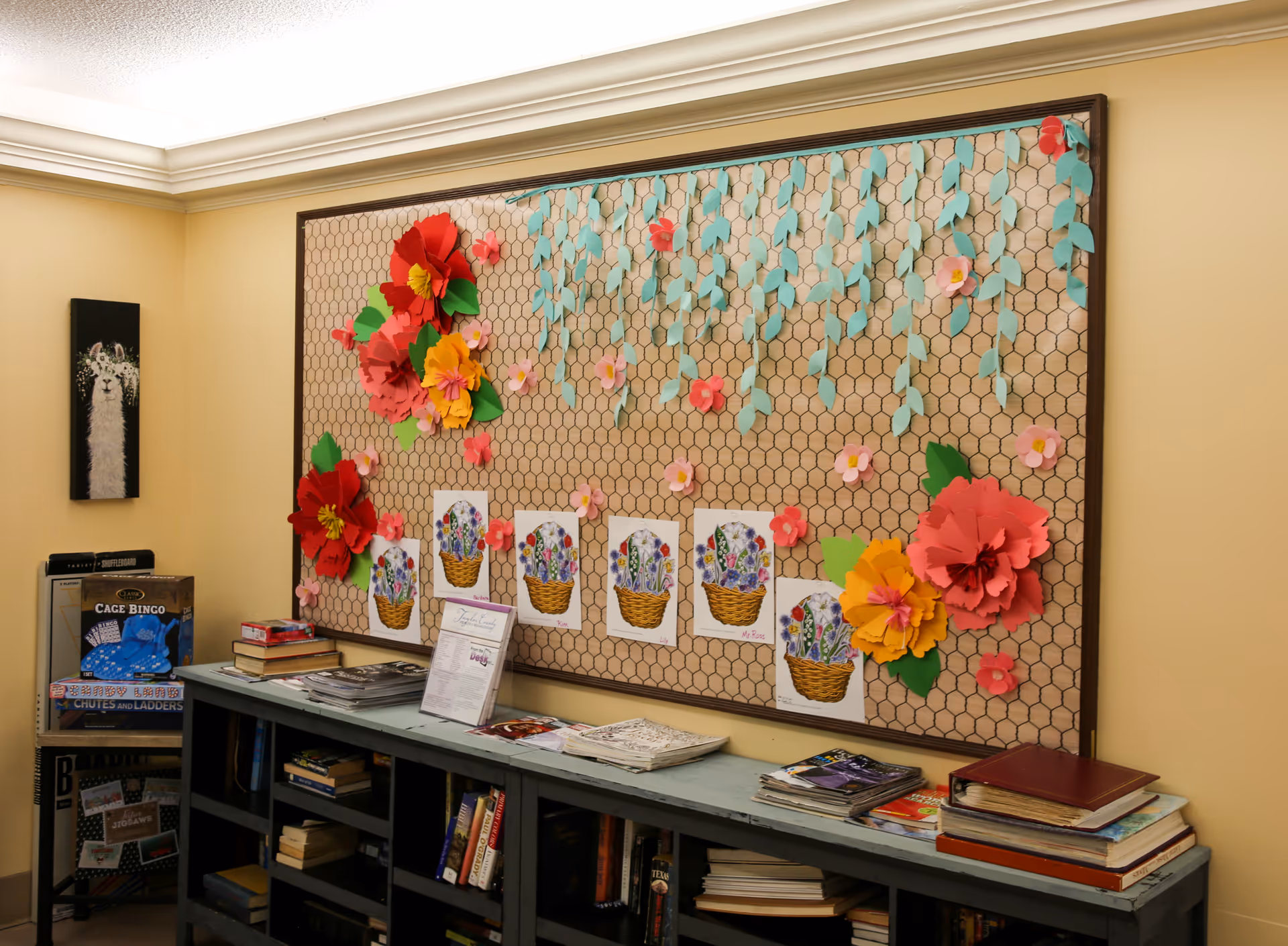 A cozy room corner with a large bulletin board decorated with colorful paper flowers and drawings of flower baskets. Below the board is a blue bookshelf filled with books and magazines. On the left side, there is a small shelf holding board games including Cage Bingo and Chutes and Ladders. The walls are painted a warm beige color.