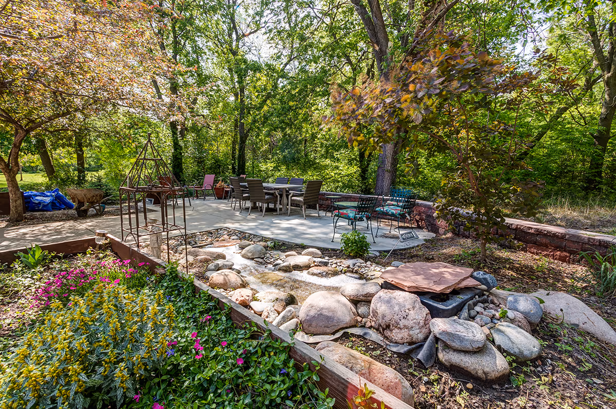 A peaceful outdoor garden area with a small rock-lined water feature in the foreground, surrounded by colorful flowers and greenery. In the background, there is a concrete patio with several chairs and tables, shaded by tall trees and lush foliage.