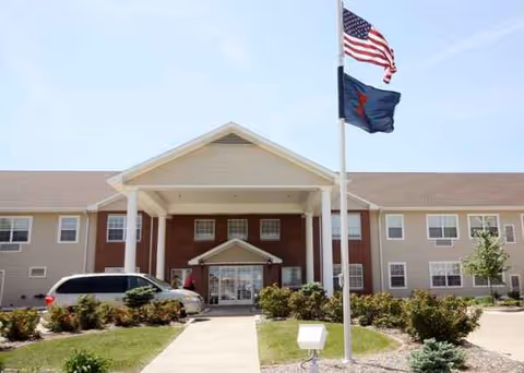 Senior living building front entrance with flags, parking area, and landscaped walkway.