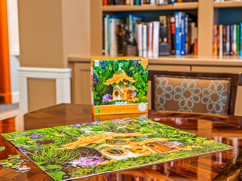 A partially completed colorful puzzle and its box sit on a polished wooden table in a room with bookshelves and a patterned chair.