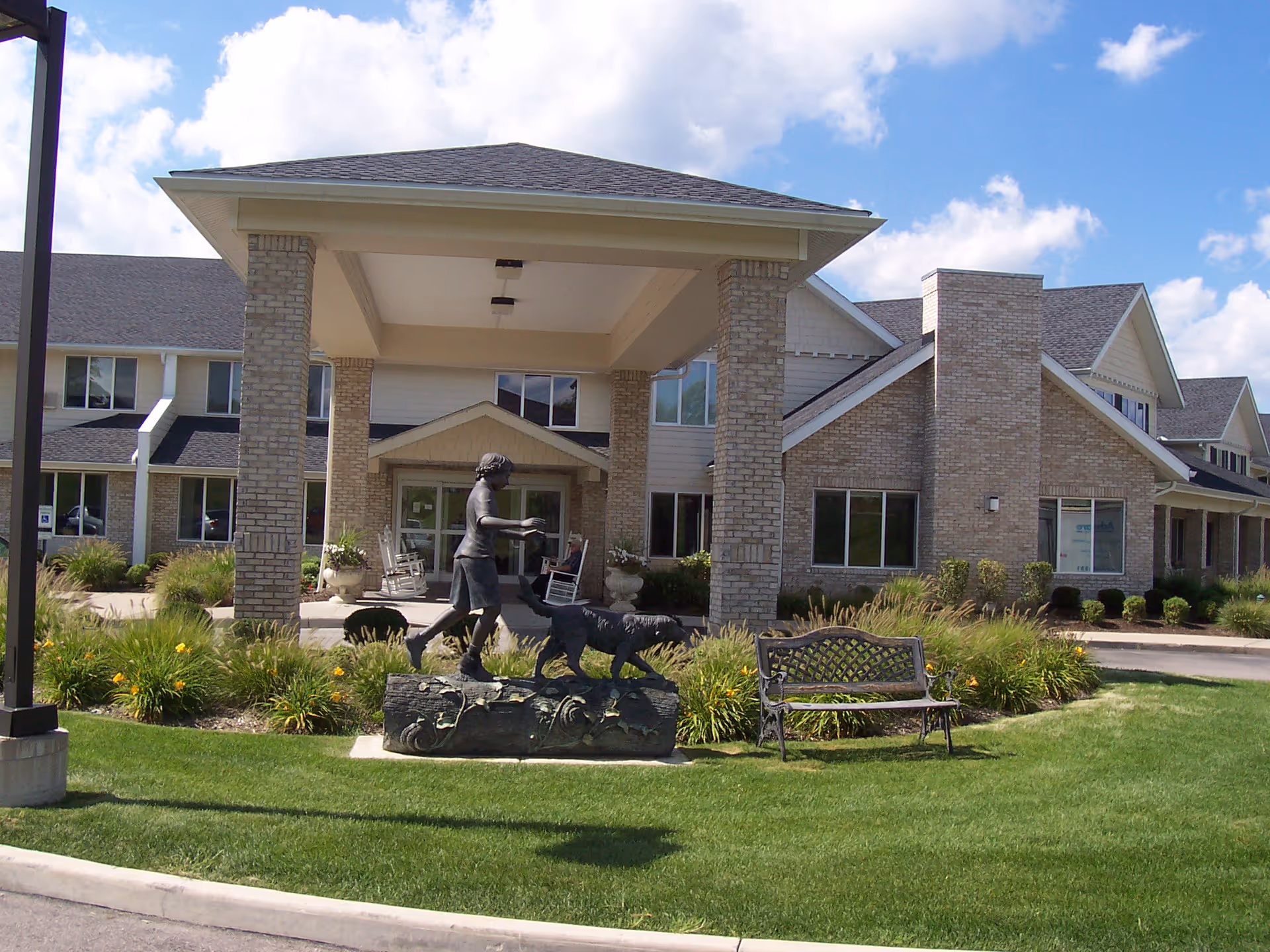 Front exterior view of Mulberry Gardens Senior Living facility with a covered entrance supported by brick columns, a bronze statue of a child walking a dog on a log, a bench, and well-maintained landscaping with green grass and flowering plants under a partly cloudy sky.