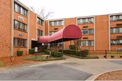 Exterior view of a multi-story brick apartment building with a red canopy over the entrance. There are windows with air conditioning units and some landscaping with bushes and mulch around the entrance area.