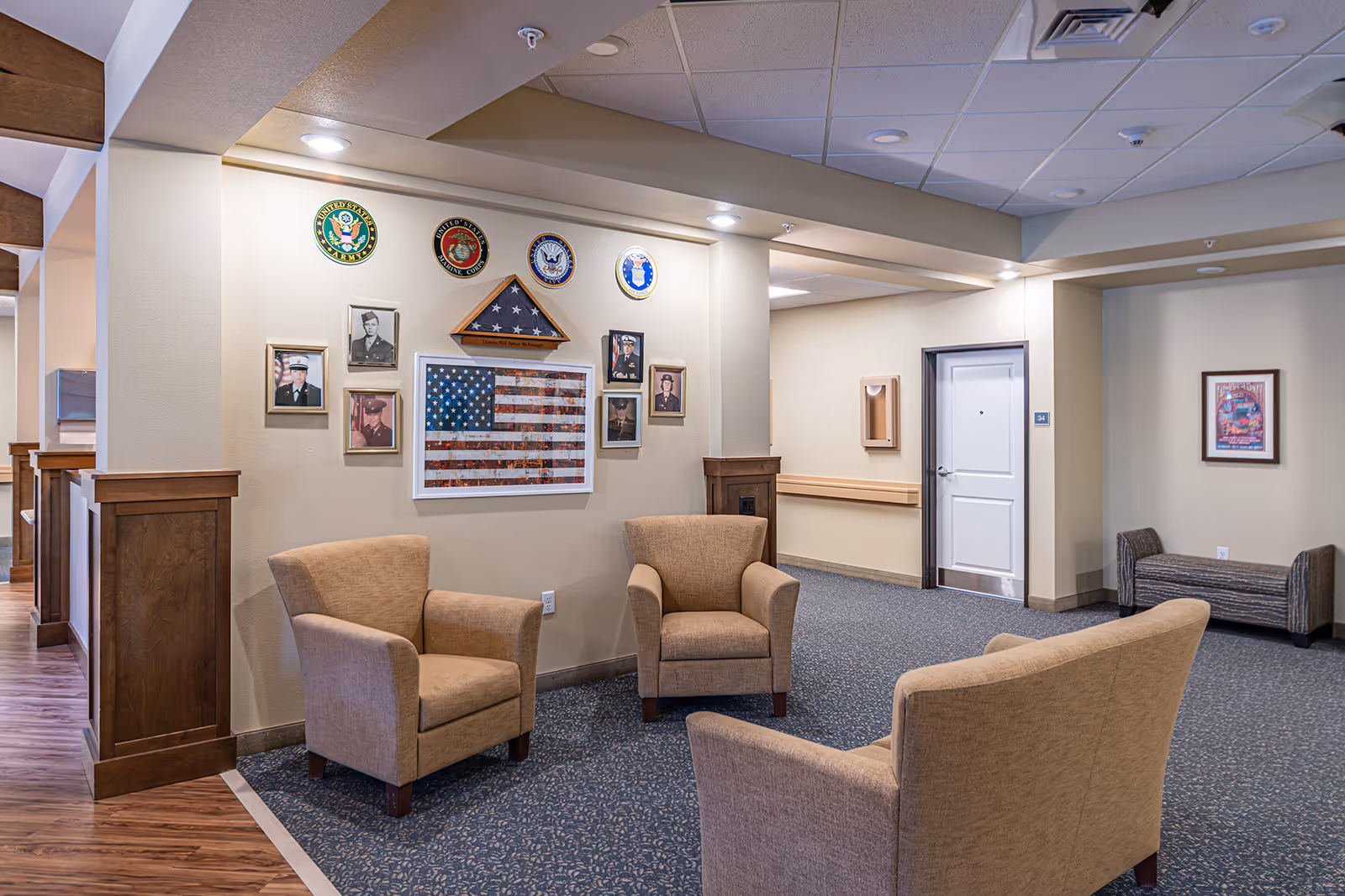 A seating area in a memory care facility with three beige armchairs arranged on a patterned carpet. The wall behind the chairs displays framed military photos, a folded American flag in a triangular case, and four military branch emblems. The space has beige walls, wood paneling, and a white door labeled with the number 34.