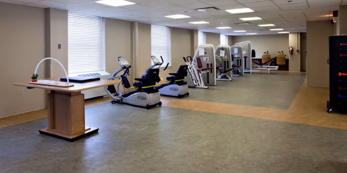 Large, well-lit exercise room with recumbent bikes, strength machines and a table in a senior living facility.