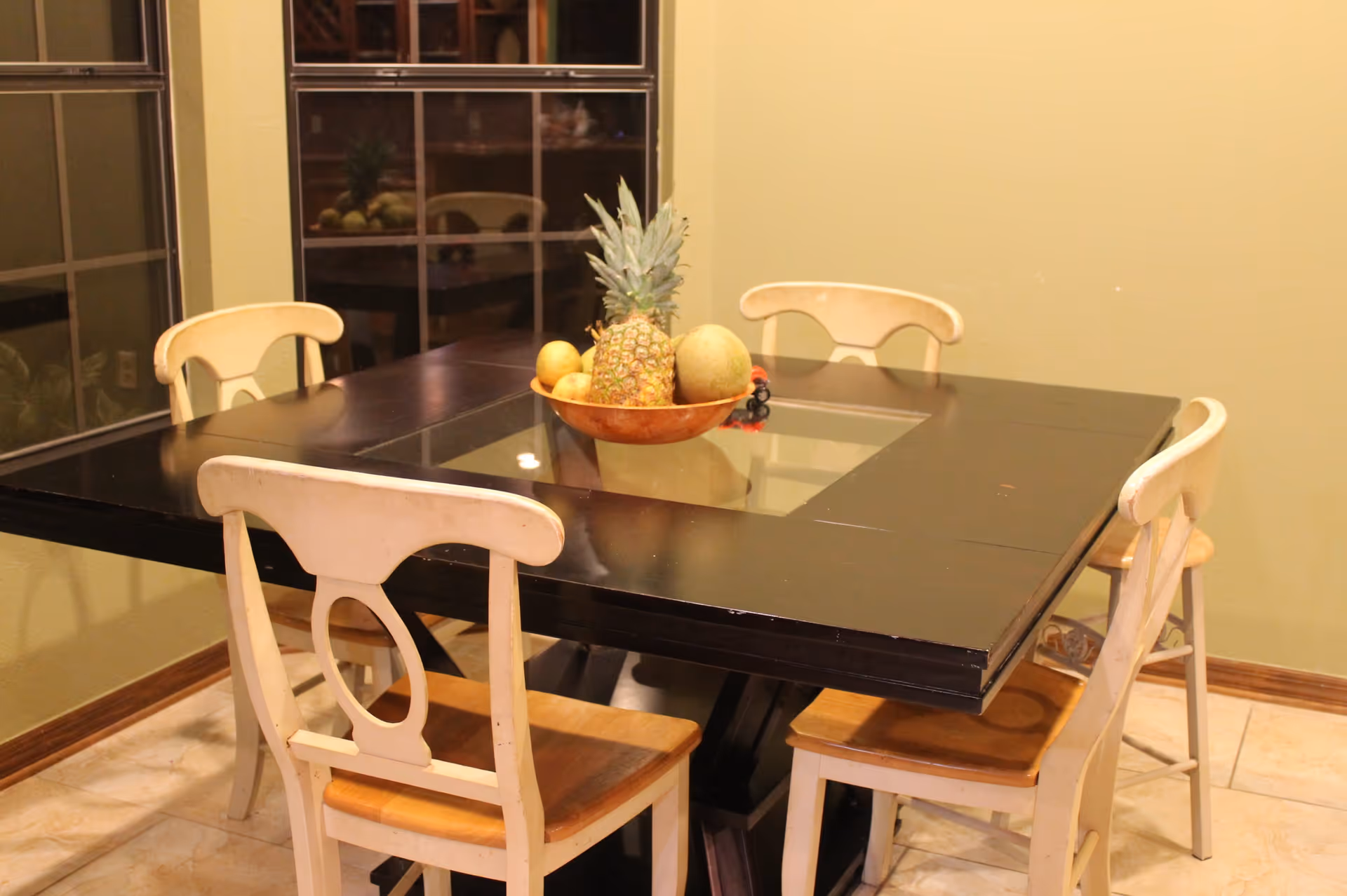 Dining room with a black wooden table, four chairs and a fruit bowl in the center near windows.