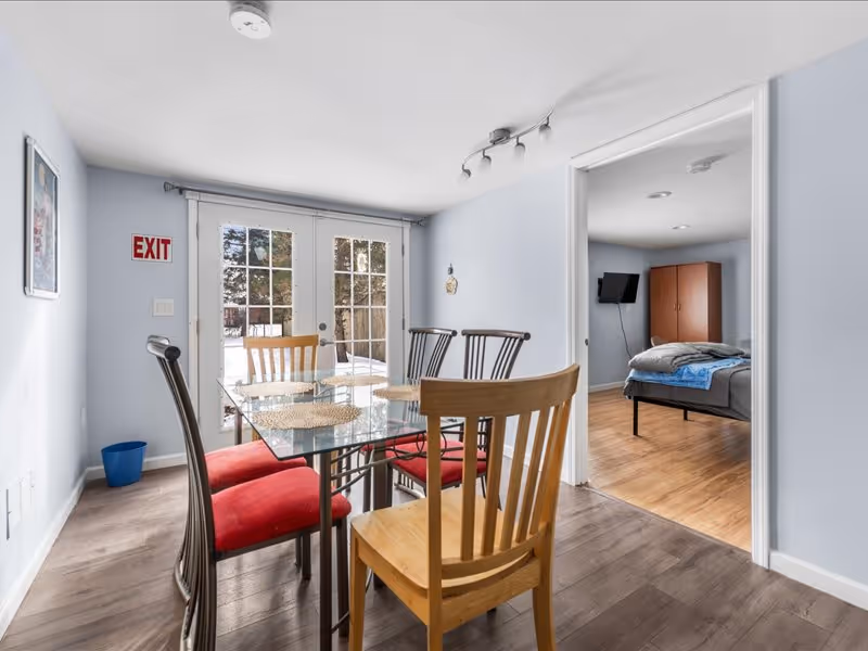 A dining area with a glass table and six chairs, four with red cushions and two wooden chairs without cushions. The room has light blue walls and wood flooring. French doors with glass panes lead outside, and an exit sign is visible on the wall. Through an open doorway, a bedroom with a bed, a TV mounted on the wall, and a wooden wardrobe can be seen.