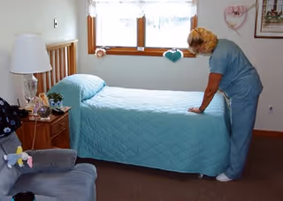 A caregiver in blue scrubs is making a bed with a light blue quilt in a senior living bedroom. The room has a wooden bed frame, a nightstand with a lamp and decorative items, a window with curtains, and a comfortable armchair with a stuffed animal on it.