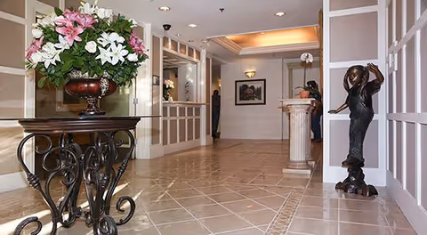 Interior view of a hallway with tiled floor, decorative wall paneling, a large floral arrangement on an ornate metal table, a bronze statue of a girl holding a hat, and a pedestal with a flower vase. The hallway is well-lit with ceiling lights and has framed artwork on the walls.