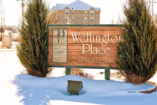 A wooden sign reading "Wellington Place 615 Hilldale Drive" stands in snow with evergreen shrubs and a multi-story building in the background.