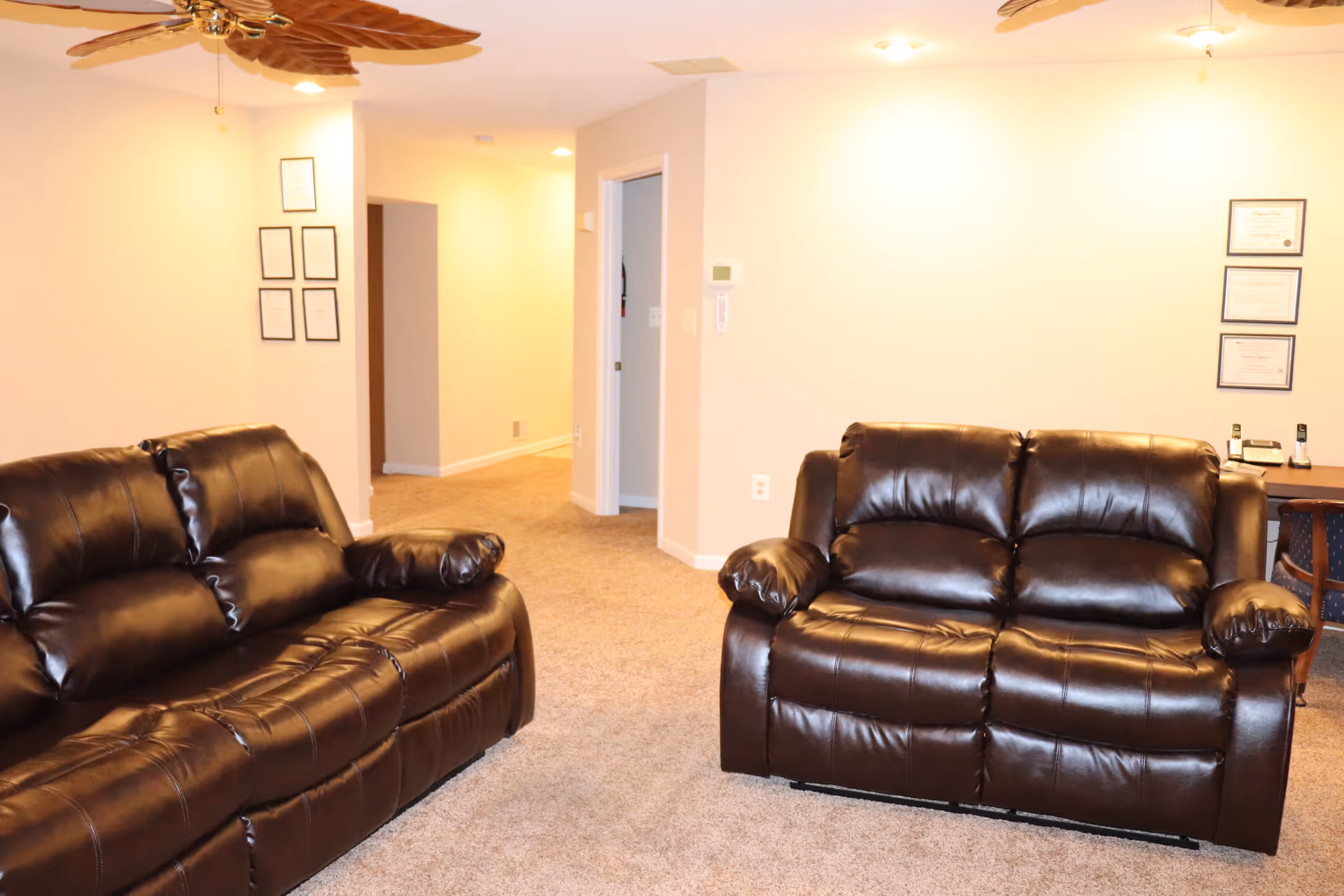 A living room with two dark brown leather sofas facing each other on a beige carpet. The walls are light-colored with framed certificates hanging on them. There is a ceiling fan with wooden blades and a hallway leading to other rooms in the background.