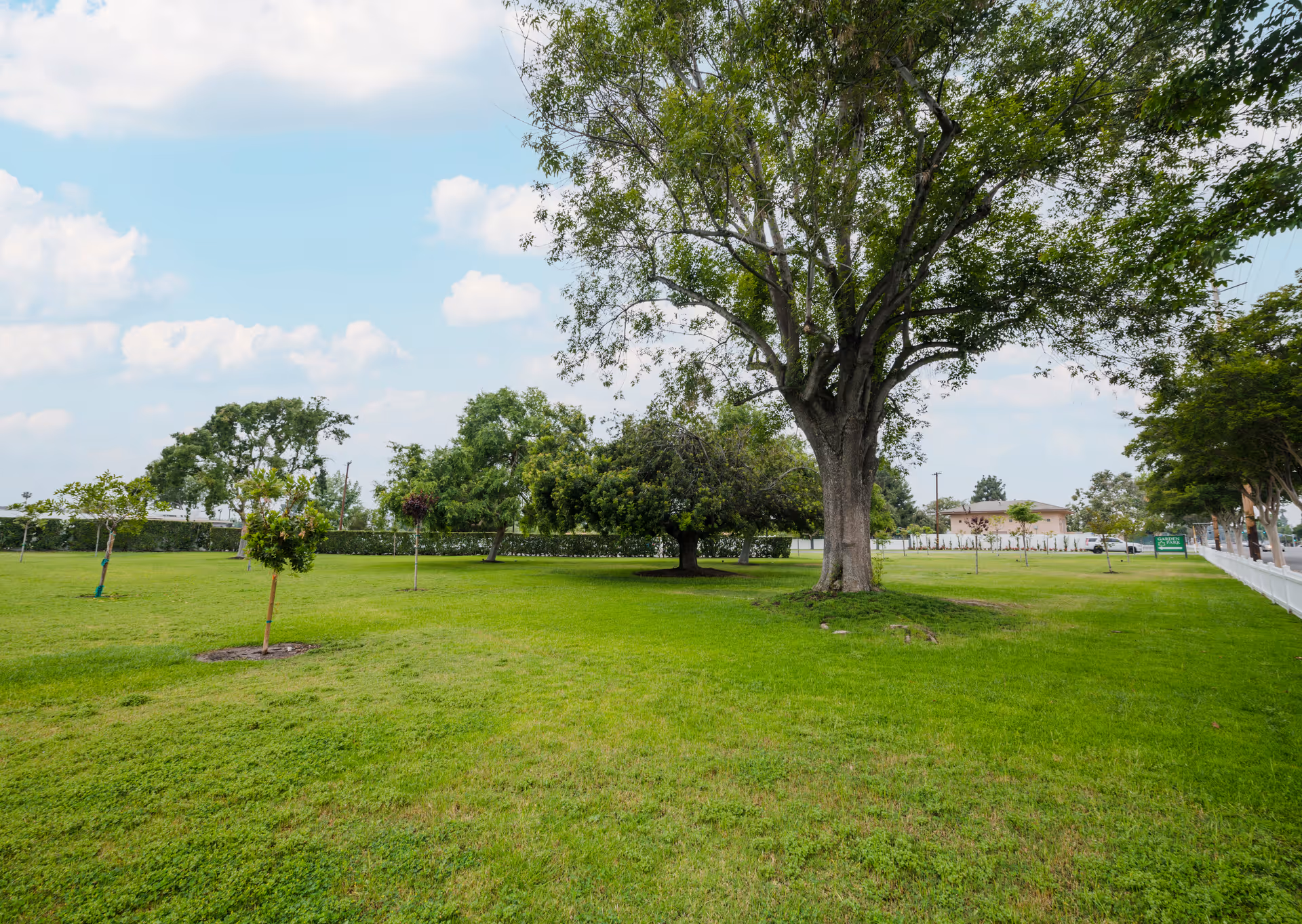 A wide grassy yard with several trees and a white fence under a partly cloudy sky.