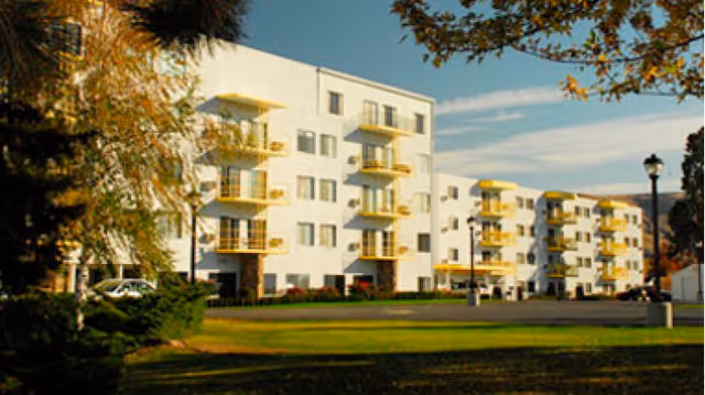 Exterior view of a multi-story senior living facility building with white walls and yellow balconies, surrounded by green grass and trees under a partly cloudy sky.