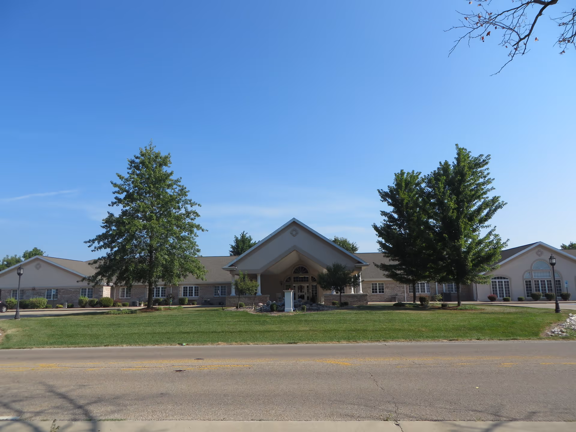 Front exterior view of a single-story senior living facility building with a peaked entrance, surrounded by green grass, trees, and a clear blue sky.