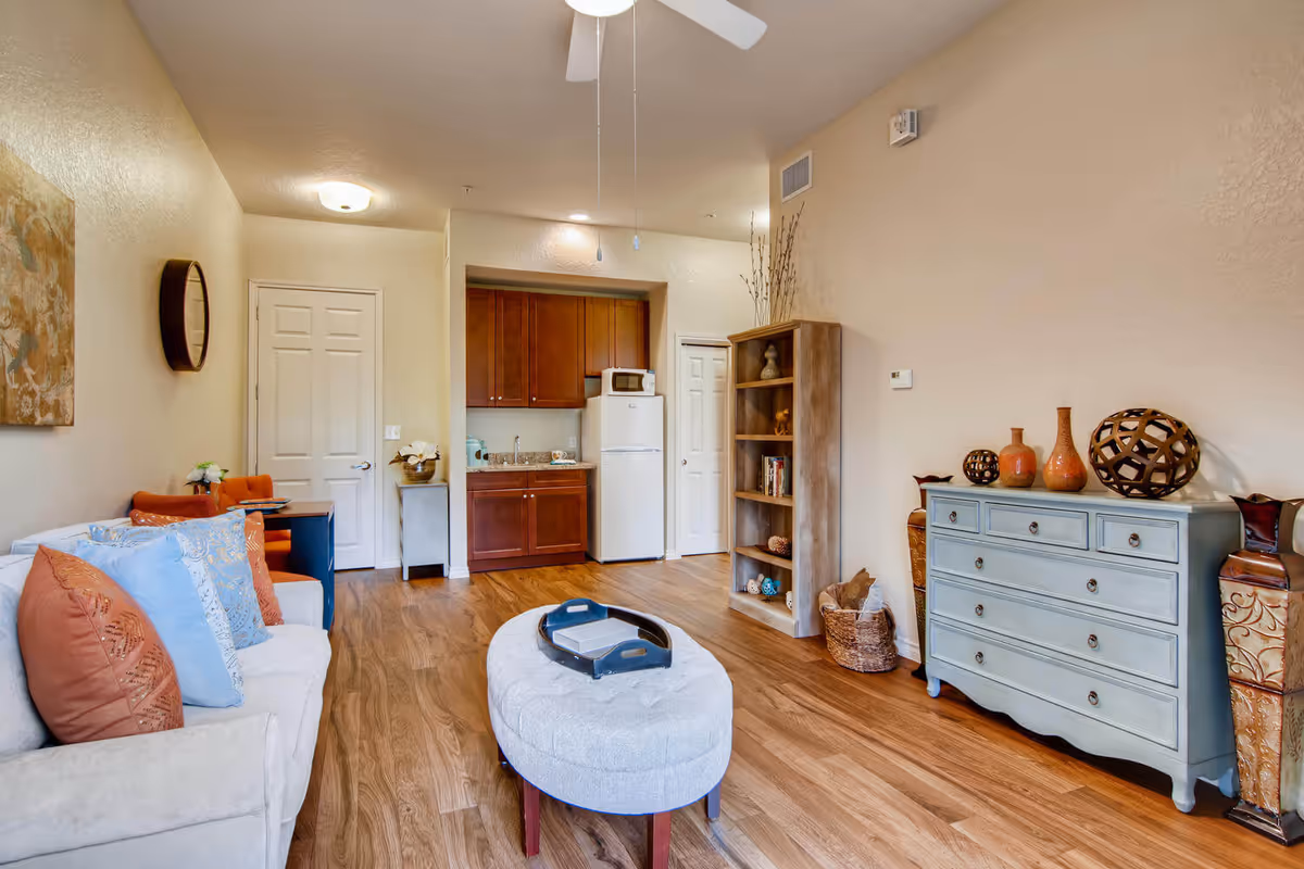 Open living room with a sofa, ottoman, bookshelf and dresser facing a small kitchenette with a refrigerator and cabinets.