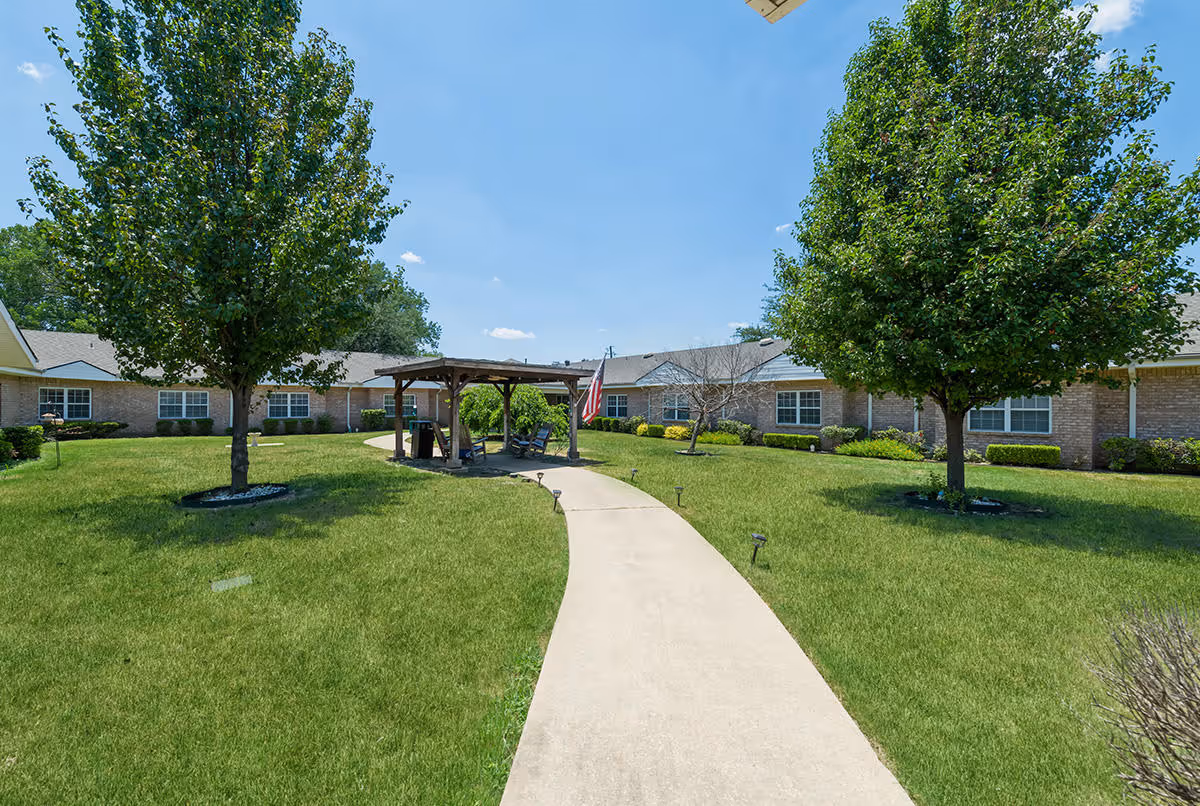 A paved walkway leads through a grassy courtyard with two large trees on either side. In the center is a wooden gazebo with an American flag hanging from it. Surrounding the courtyard are single-story brick buildings with multiple windows under a clear blue sky.