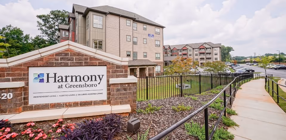 Exterior view of Harmony at Greensboro senior living facility showing a large brick sign with the facility name and services offered. The multi-story building is visible in the background along with a parking lot, landscaped garden, and a paved walkway with a black metal railing.