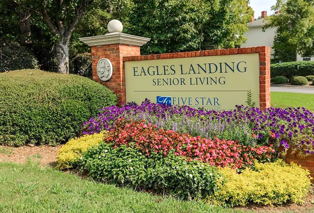 A landscaped entrance sign for Eagles Landing Senior Living, surrounded by colorful flowers and green bushes, with trees and a building in the background.
