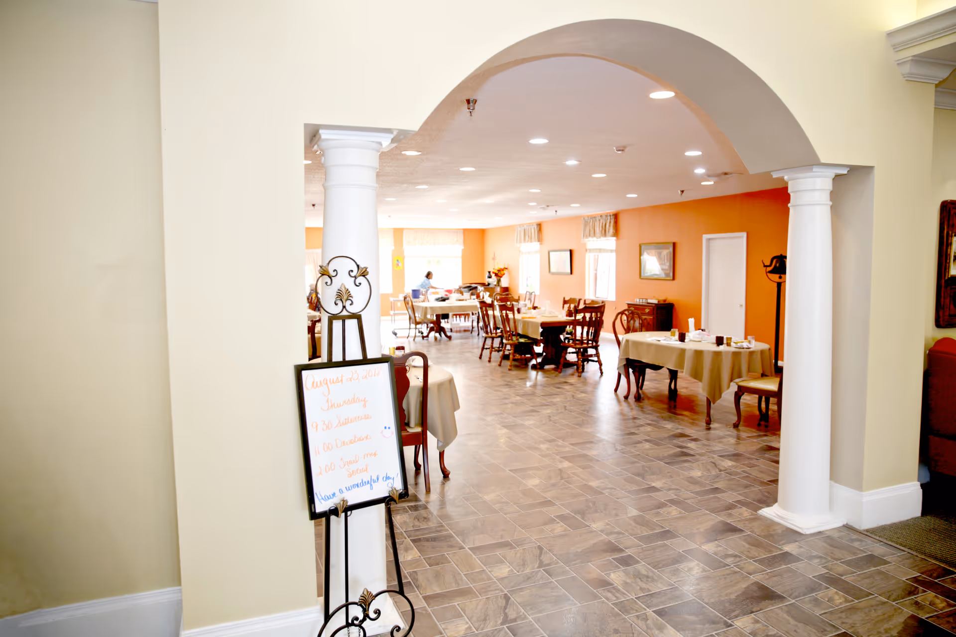 View through an archway into a dining room with several tables and chairs arranged for meals. The room has orange walls, framed pictures, and windows with curtains letting in natural light. A whiteboard with a schedule is placed near the archway, and a person is seated at a table in the background.