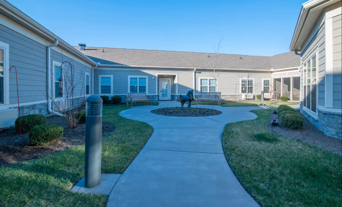 Outdoor courtyard area at Morning Pointe of Lexington featuring a concrete walkway leading to a circular garden bed with a horse statue in the center, surrounded by benches, grass, shrubs, and a clear blue sky.
