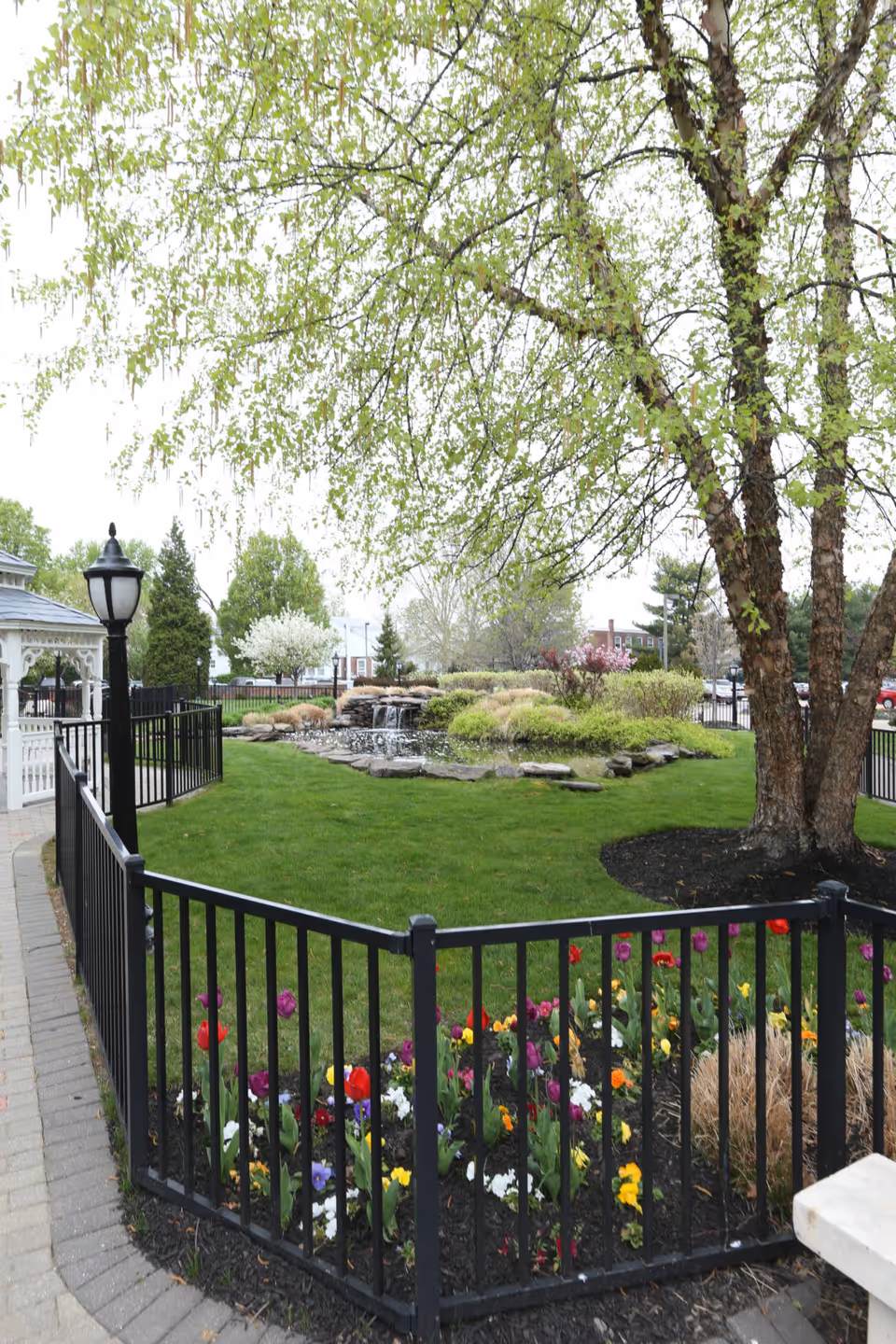 A well-maintained outdoor garden area with a black metal fence surrounding colorful flowers. There is a large tree with green leaves, a small waterfall feature with rocks, and a white gazebo visible on the left side. The background includes some trees, shrubs, and a few buildings.