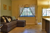 A cozy living room area with a dark gray sectional sofa adorned with multiple cushions. The room has beige walls and tiled flooring. Two decorative wall hangings are visible above the sofa. A window with sheer white curtains allows natural light to enter. Adjacent to the living room is a dining area with a hanging light fixture and similar sheer curtains on the window.