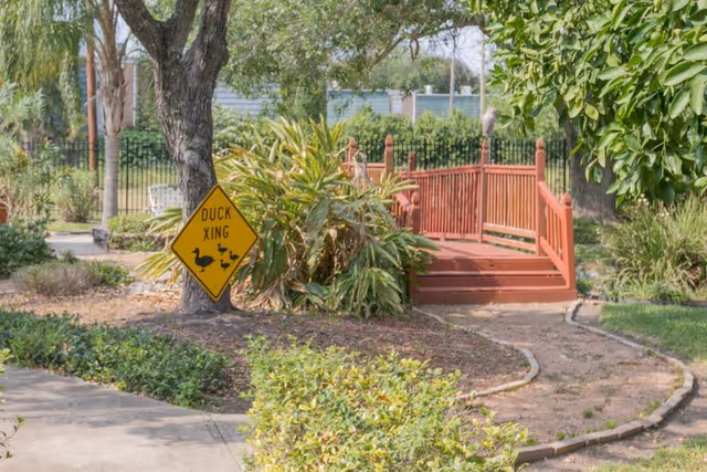 A garden area with a small red wooden bridge over a dry path, surrounded by green bushes and trees. A yellow diamond-shaped sign near a tree reads 'DUCK XING' with images of ducks crossing.