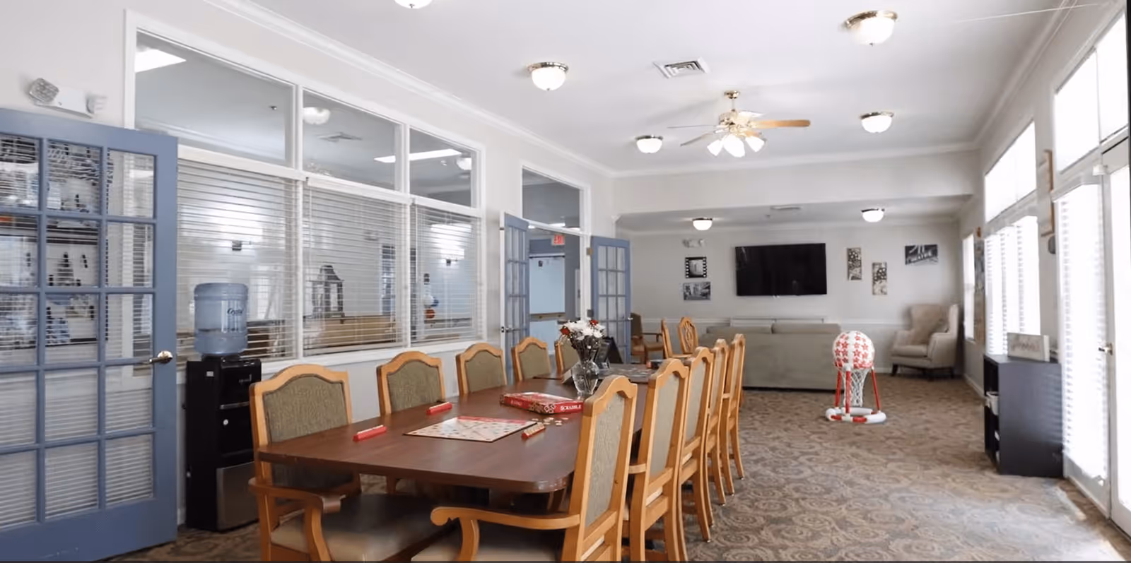 A spacious common area in an assisted living facility featuring a long wooden table with chairs arranged around it. On the table are a Scrabble board game and a vase with flowers. The room has carpeted floors, multiple ceiling lights, a ceiling fan, and large windows with blinds letting in natural light. In the background, there is a seating area with a couch, armchair, wall-mounted TV, and decorative wall art. Double glass doors and a water cooler are visible on the left side.