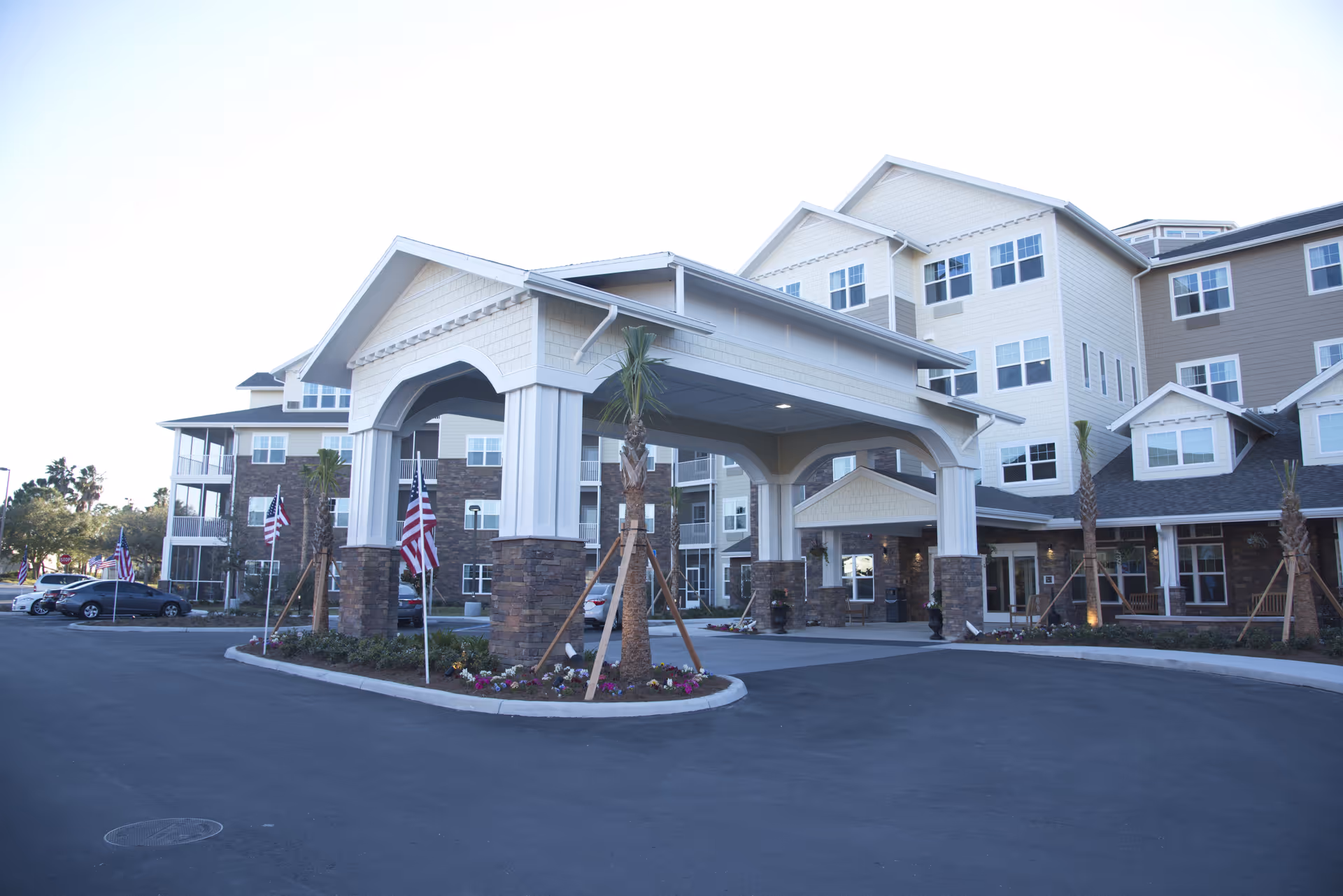 Multi-story senior living building with a covered porte-cochère entrance, flagpoles, landscaping, and parked cars.