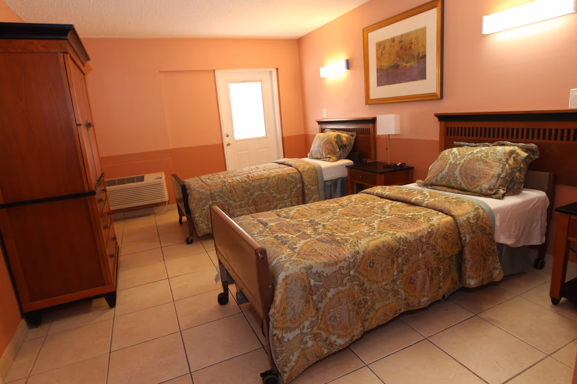A bedroom with two single beds, each with patterned bedspreads and pillows. There is a wooden nightstand with a lamp between the beds, a wooden armoire on the left, and a framed artwork on the wall above the beds. The room has peach-colored walls and tiled flooring, with a door at the far end.