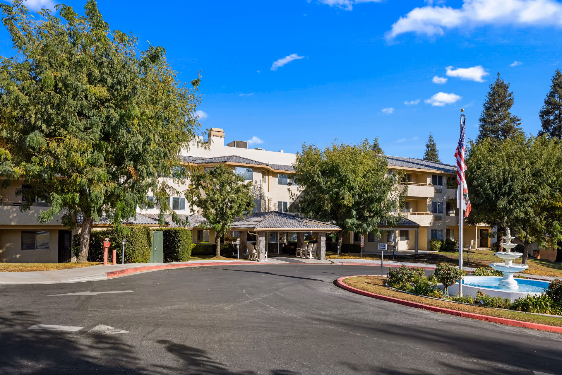 Exterior view of Columbus Estates - A Provincial Senior Living Community showing a three-story building surrounded by trees, a driveway with a covered entrance, an American flag on a flagpole, and a white fountain in a landscaped area under a blue sky with some clouds.