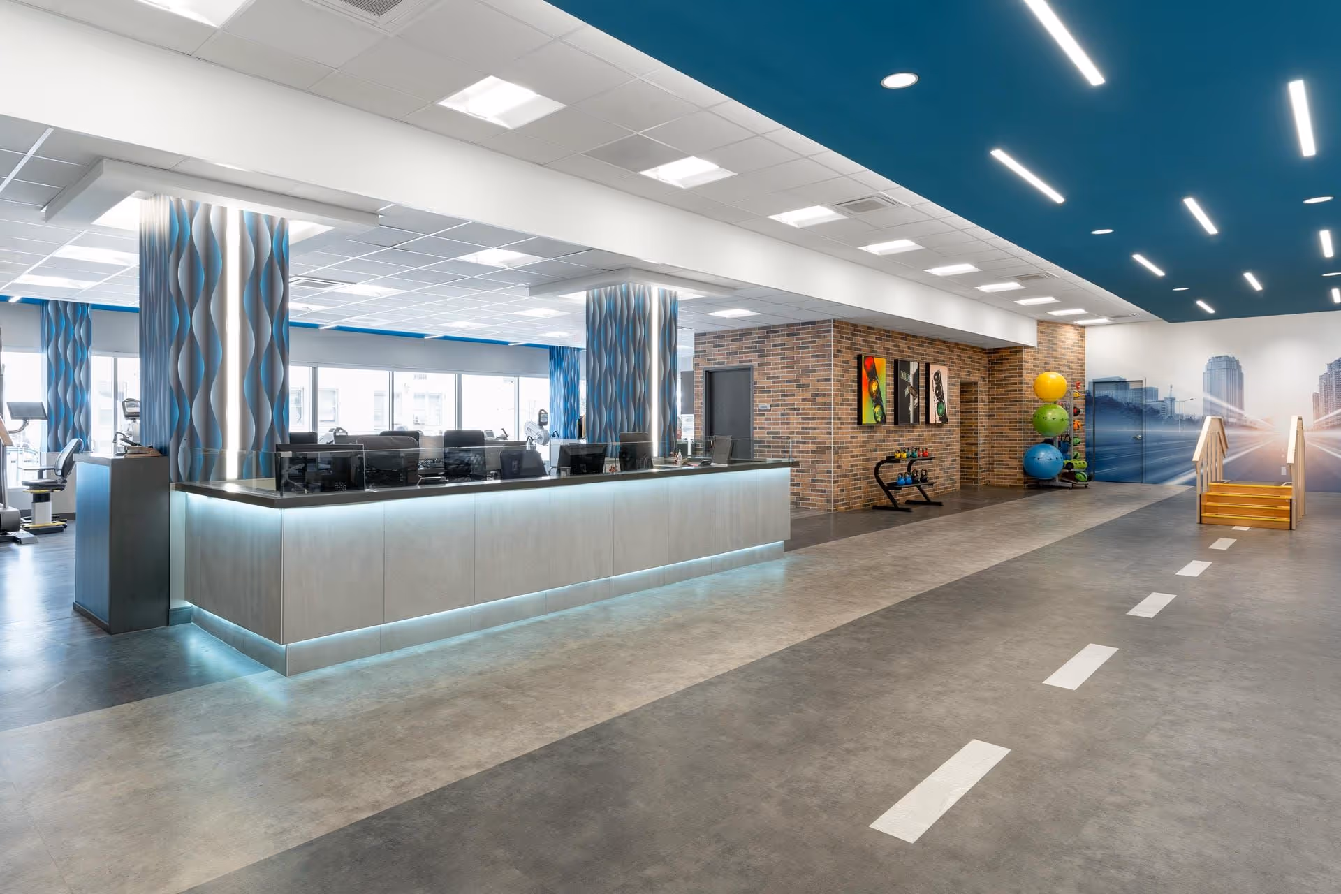 Spacious modern interior with a long illuminated reception desk, exercise/therapy equipment, and a marked corridor.