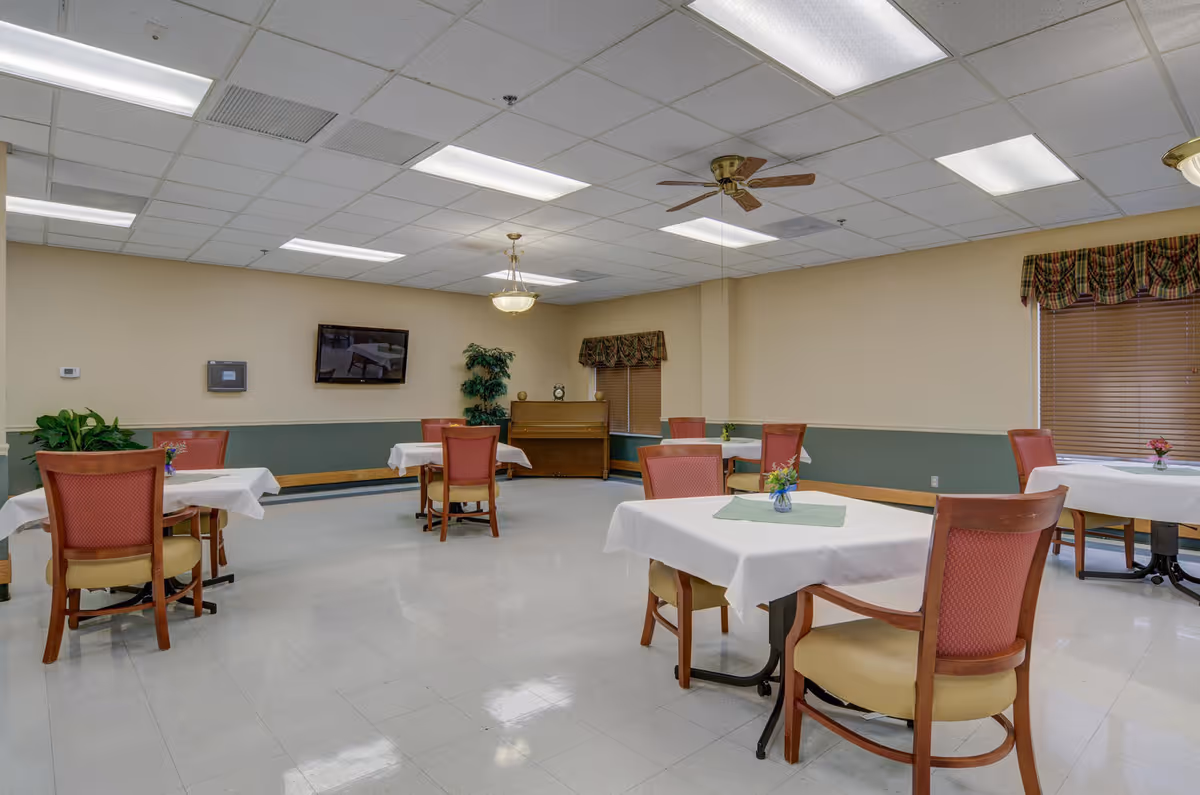 A dining room in a senior living facility with several tables covered with white tablecloths and small flower vases. The room has beige walls with a green lower half, a ceiling with fluorescent lights and ceiling fans, a wall-mounted TV, a piano, and windows with plaid valances and blinds.
