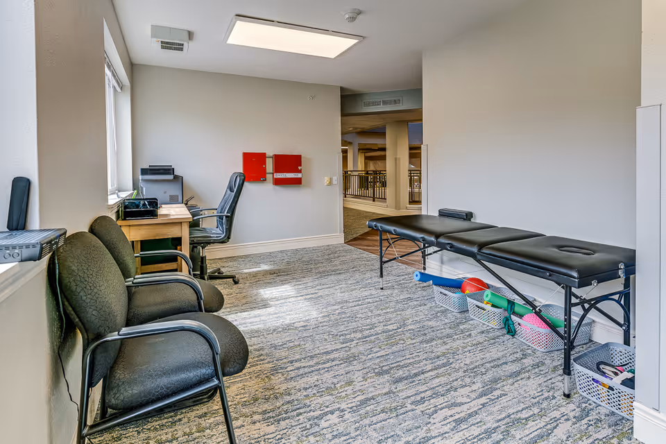 A small interior therapy room with a black portable massage table, three guest chairs along the wall, and a desk with an office chair and printer.