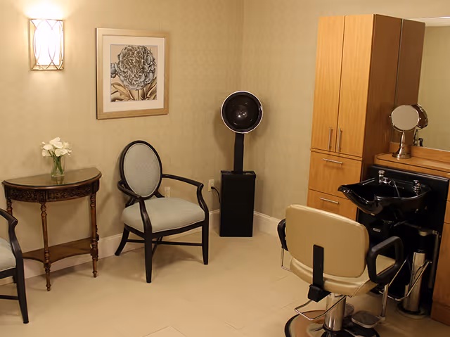Small salon-style interior with a styling chair and sink, hooded hair dryer, waiting chairs, side table, and storage cabinet.