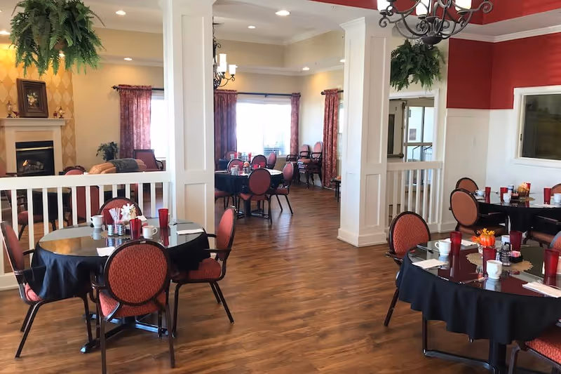 A dining room in a senior living facility with round tables covered in black tablecloths, red cushioned chairs, and place settings including cups and red glasses. The room has wooden flooring, large windows with red curtains, a fireplace, and decorative plants hanging from the ceiling.
