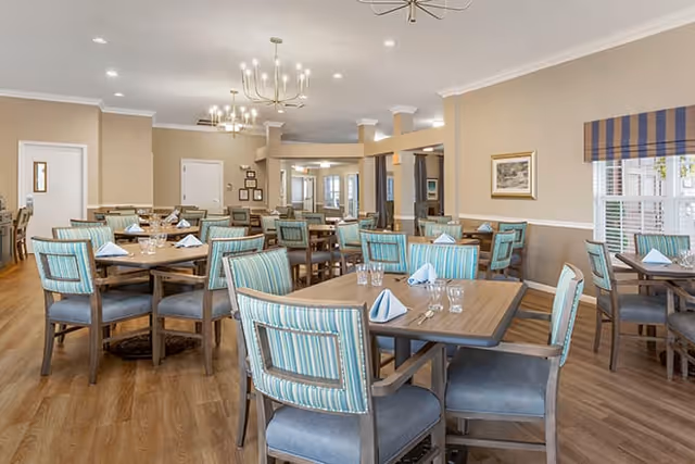 A spacious dining room with multiple wooden tables and chairs featuring blue and green striped upholstery. Each table is set with folded napkins, glasses, and silverware. The room has light beige walls, wooden flooring, and modern chandeliers hanging from the ceiling. Windows with striped valances allow natural light to enter the space.