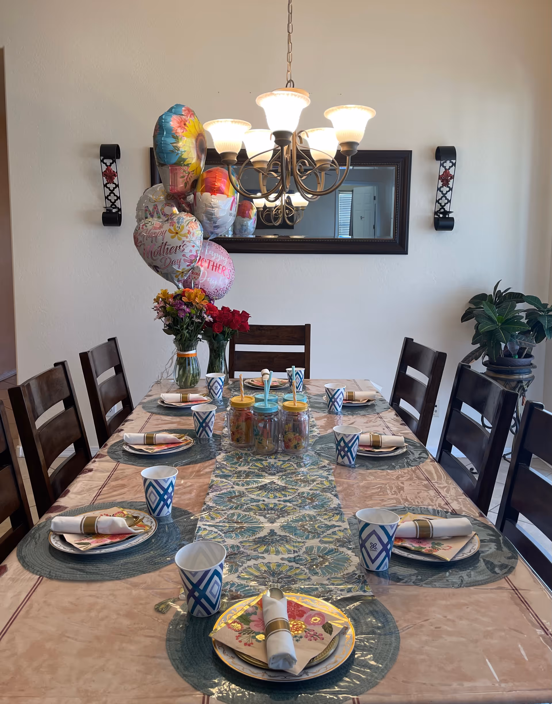 A dining table set for a meal with eight place settings, each with a plate, napkin, and paper cup. The table has a decorative table runner and placemats. There are two vases with flowers and several colorful balloons tied to one vase. A chandelier with five lights hangs above the table, and a large mirror is mounted on the wall behind it. Two wall sconces flank the mirror, and a potted plant is visible in the corner.