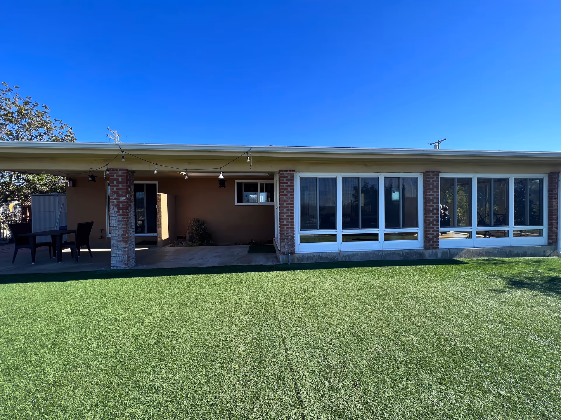 Single-story building exterior with a covered patio supported by brick columns, large windows, string lights and a green lawn in front.
