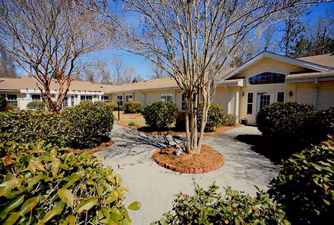 Outdoor courtyard area of a senior living facility with a paved walkway, trimmed bushes, and leafless trees under a clear blue sky. The building surrounding the courtyard is single-story with light-colored siding and multiple windows.