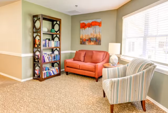 Small seating area with a red loveseat, striped armchair, bookshelf, lamp and a window in a common room.