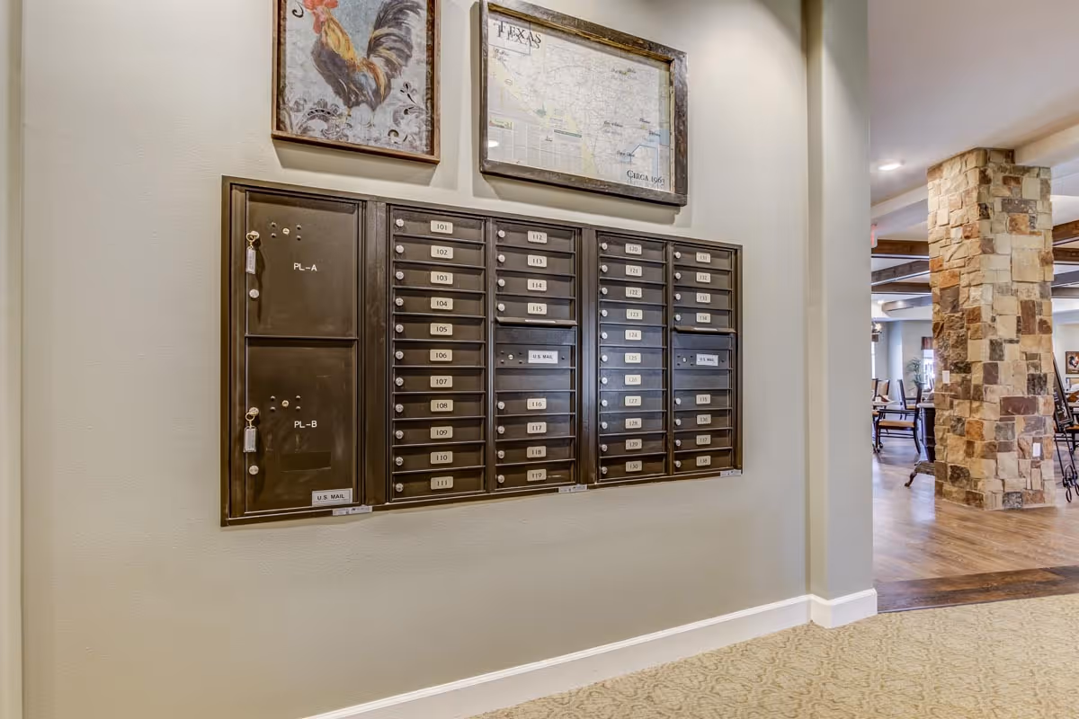 Wall-mounted mailboxes with numbered slots and two locked compartments labeled PL-A and PL-B, set against a beige wall. Above the mailboxes are two framed pictures, one of a rooster and the other a map of Texas. To the right, a stone pillar and part of a dining area with wooden chairs and tables are visible.