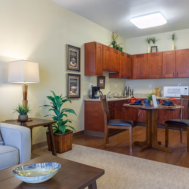 Open living area with a kitchenette of wood cabinets, a round dining table set, and a sofa with lamp and potted plants.