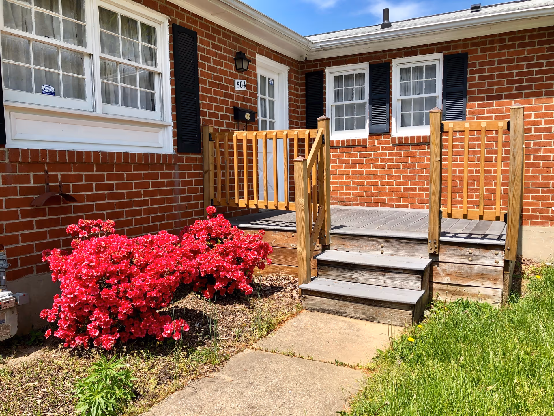 Front entrance of a brick building with white-framed windows and black shutters. There is a small wooden porch with steps and railings leading up to a white door labeled with the number 504. Bright red flowers and green grass surround the concrete walkway leading to the porch.