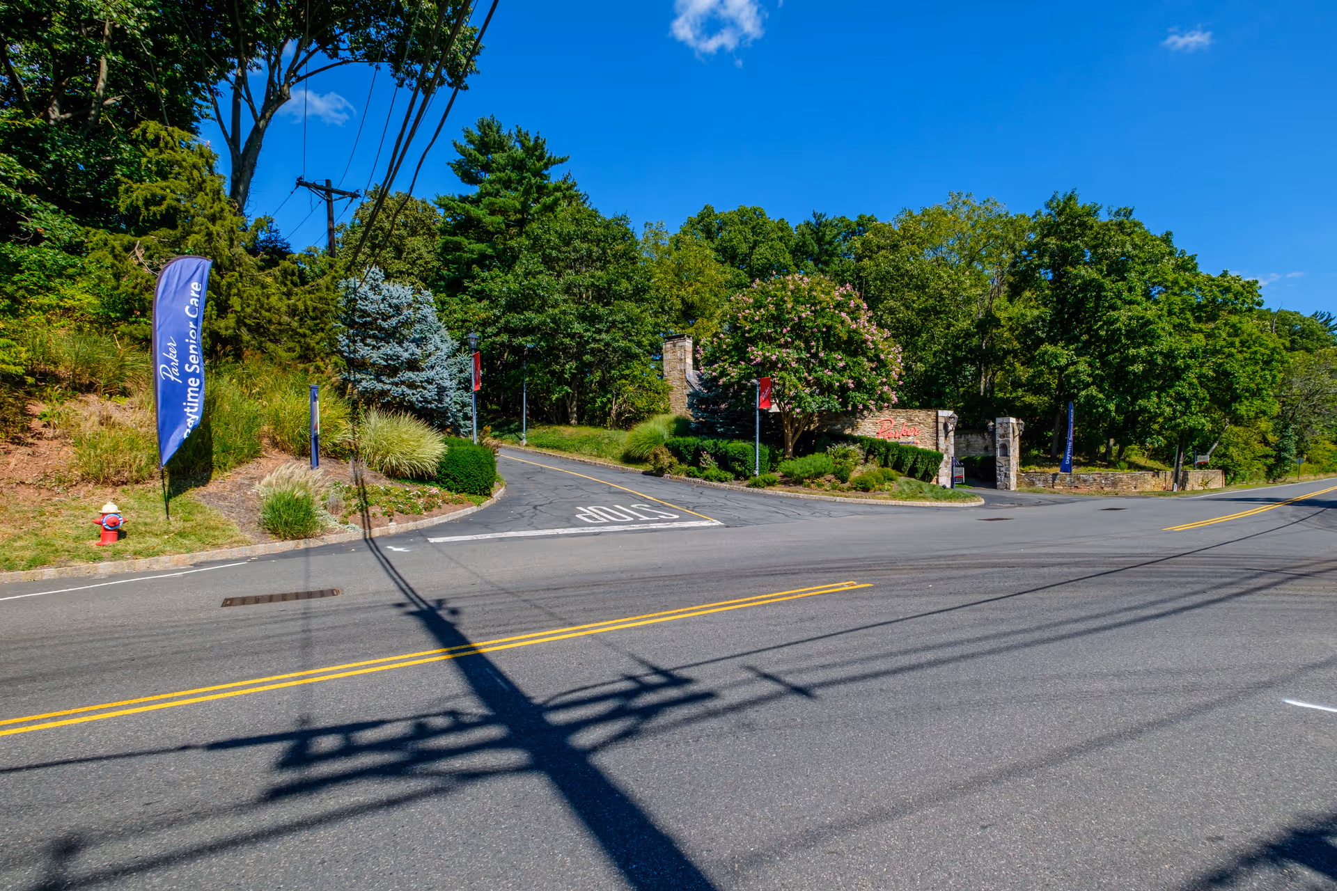 Entrance to Parker at Stonegate facility with a stone wall and archway surrounded by trees and greenery under a clear blue sky. A road with yellow dividing lines and a stop marking is in the foreground. Blue flags with the text 'Parker Daytime Senior Care' are visible near the entrance.