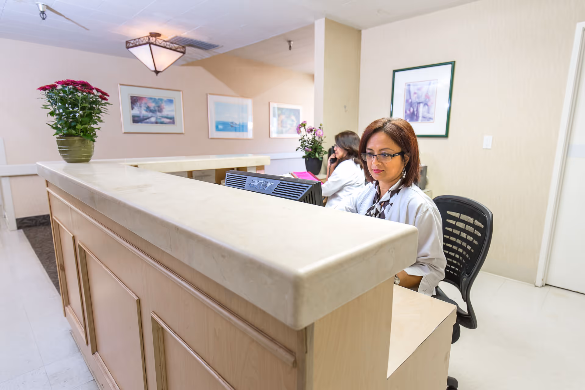 Reception area with two women working behind a beige marble counter. One woman is focused on a computer while the other is on the phone. The walls are decorated with framed pictures and there are potted plants on the counter and in the background.